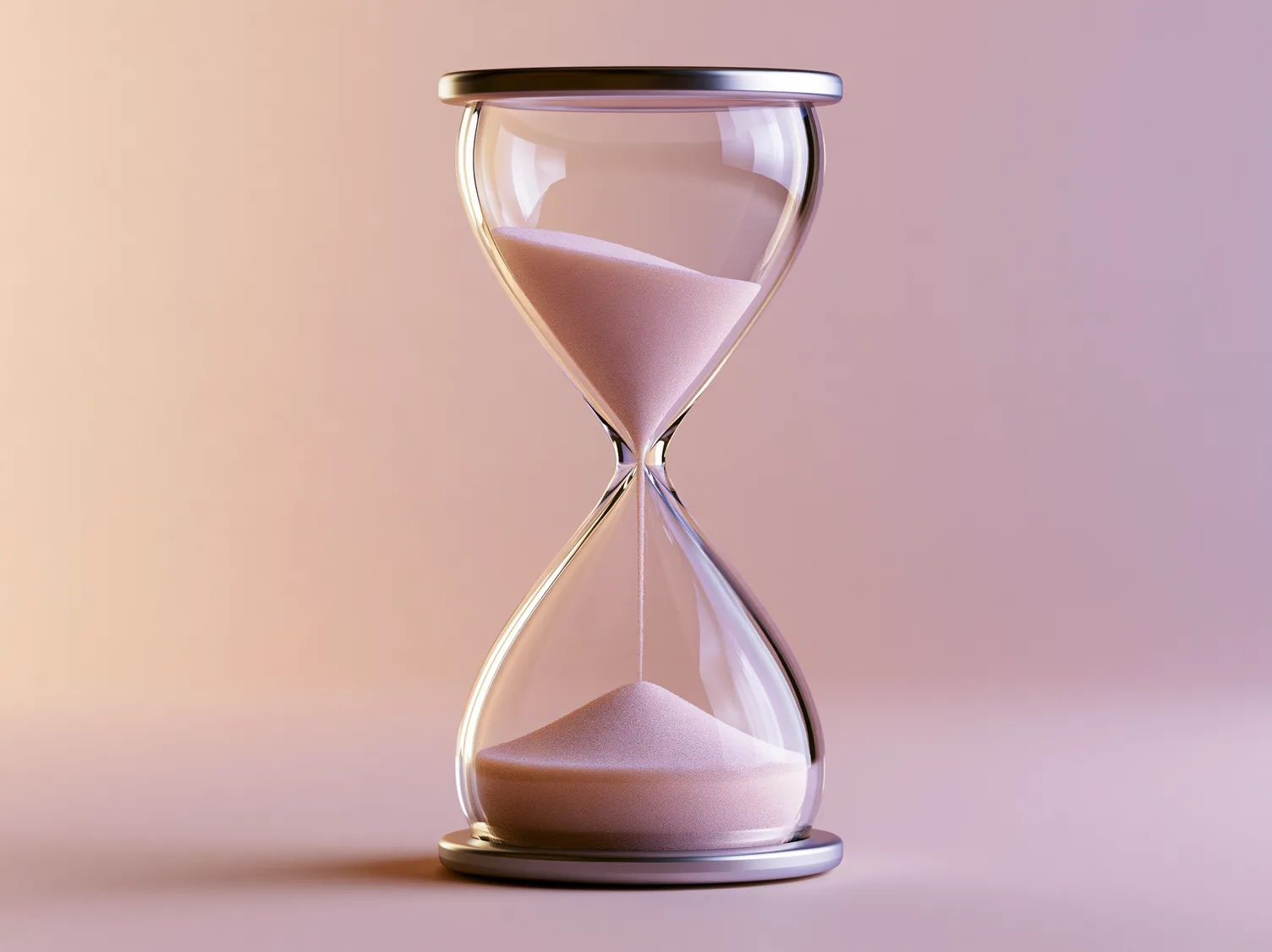 An hourglass in clear glass with brushed metal caps, pale sand heaped in both bulbs and a thin thread of it still falling between them, against a backdrop shading from peach to pink.